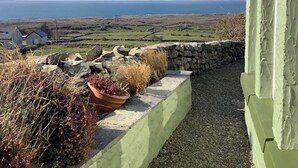 Un mur de pierres avec un pot de fleurs, une zone herbeuse et une vue sur la mer et les maisons au loin.