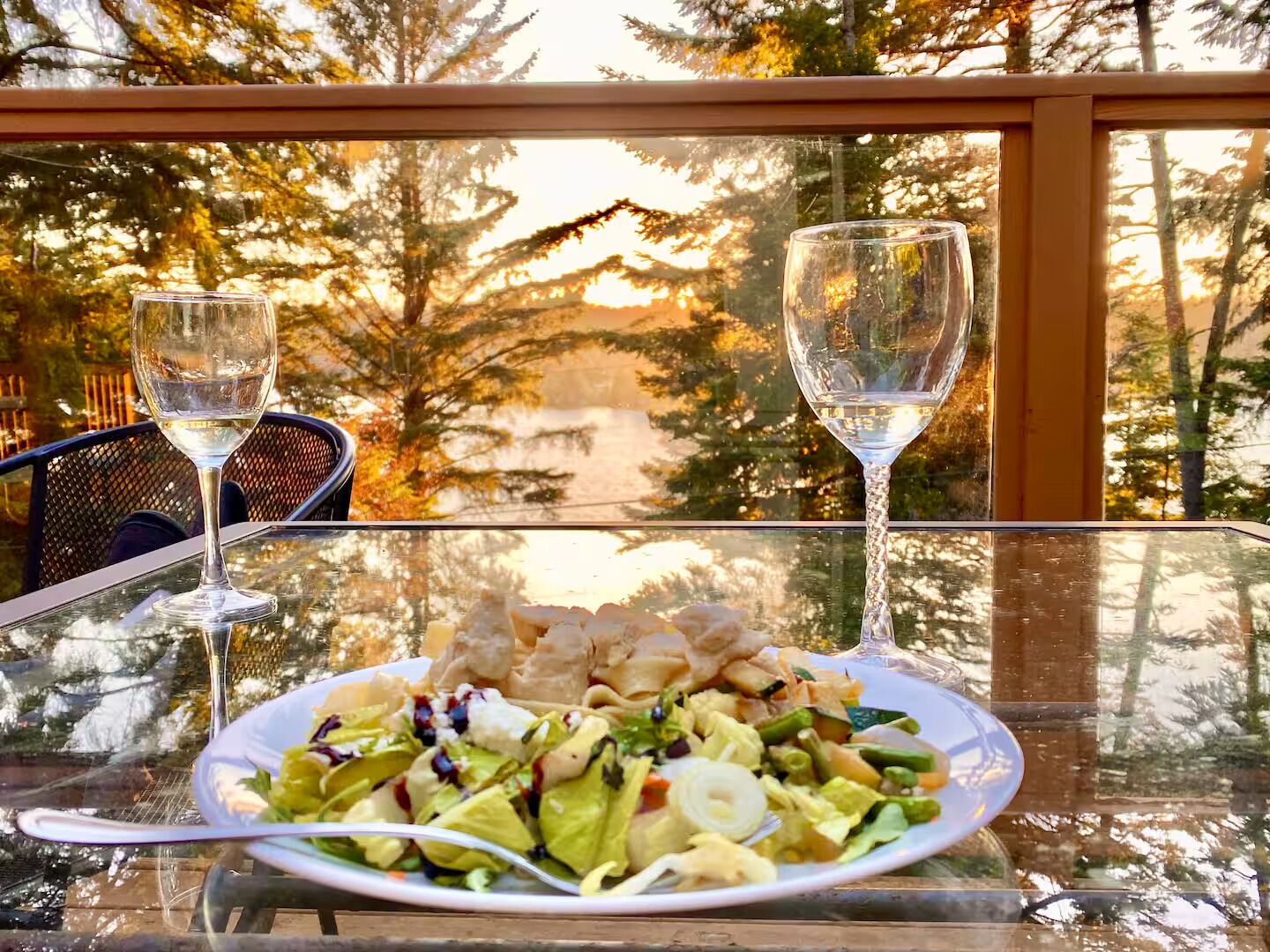 A dining table with a plate of food and two glasses of white wine, set against a backdrop of trees and a clear sky.