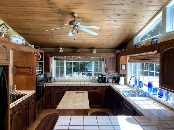 A kitchen with wooden cabinets, a central island, a ceiling fan, and a window with a view of greenery. - Private One-Acre Woahink Lake View Retreat, Hot Tub & Dock Access (FLORENCE)