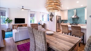 Een moderne woonkamer met een bank, eettafel en stoelen, een keuken met een roestvrijstalen koelkast, en een plafondventilator.