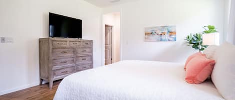A bedroom with a bed, a wooden dresser, a flat-screen TV, a painting, and a plant.