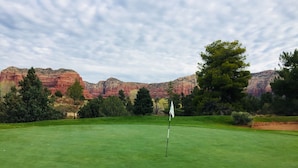 Un campo de golf con un marcador central, rodeado de hierba verde y árboles, con formaciones rocosas rojas al fondo.