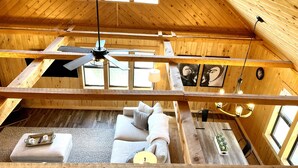A cozy cabin interior with a loft bedroom, a dining area, and a living room with a sofa and pillows.
