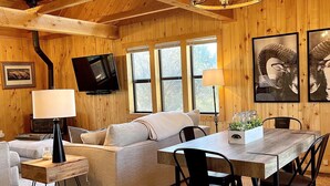 A cozy wooden interior with a sofa, dining table, chairs, a TV, and framed artwork.