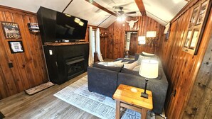 Ein gemütliches Zimmer mit Kamin, einer Couch, einem Fernseher und einem Deckenventilator.
