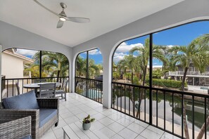 A spacious balcony with a view of a pool and palm trees, furnished with wicker chairs and a table.