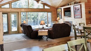 Een gezellig chaletinterieur met houten plafond, grote ramen, een open haard en een televisie.