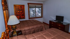 A bedroom with two beds, a television on a dresser, a window with a view of snow-covered trees, and a lamp on a nightstand.