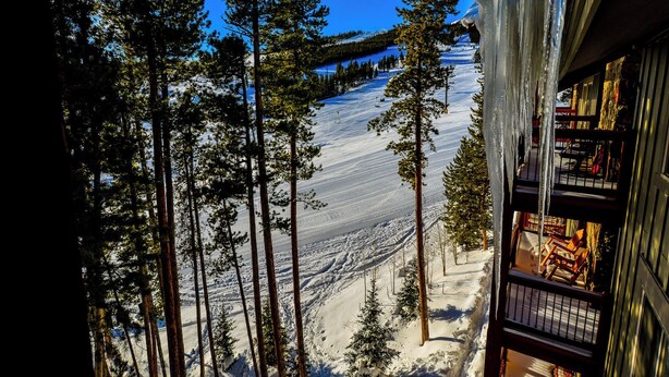 A view of a snowy mountain landscape with tall pine trees, a ski slope, and a building with a balcony overlooking the scene.