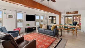 A spacious living room with a ceiling fan, a fireplace, a dining area with a wooden table and chairs, and large windows offering a view of the outdoors. - West Bend Lake House-Sleeps 12- Pontoon Avail-Full Pickleball & Basketball Court (West Bend)