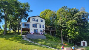 A two-story house with a deck and a dock extending into a body of water. - West Bend Lake House-Sleeps 12- Pontoon Avail-Full Pickleball & Basketball Court (West Bend)