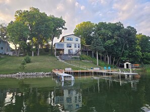 Uma casa de dois andares com um cais que se estende por um lago calmo, cercada por árvores.