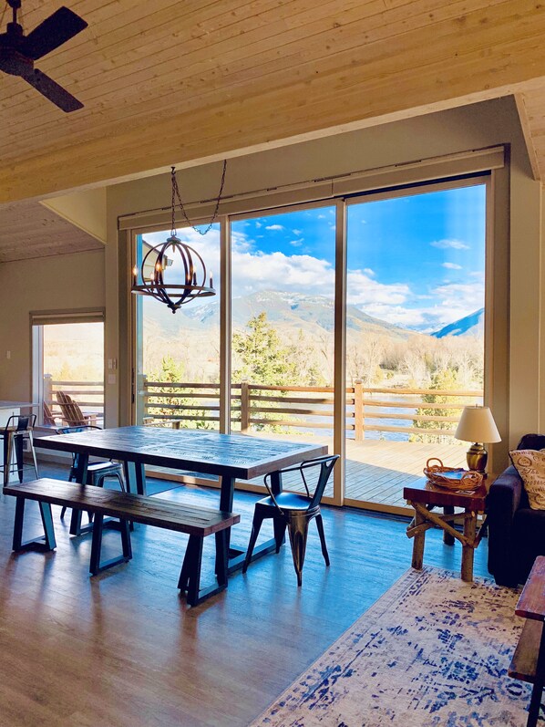 Une pièce spacieuse avec une grande table en bois, des bancs et une vue sur les montagnes, visible à travers de grandes fenêtres.