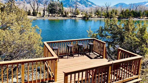 Una terraza de madera con dos mesas y sillas, con vista a un lago y montañas al fondo.