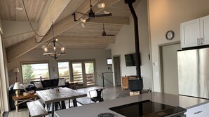 Una cocina amplia con una isla central, horno empotrado y estufa. Hay un comedor con mesa y sillas, además de un sofá. La cocina tiene vigas de madera en el techo y un ventanal grande.