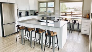 Una cocina moderna con una isla grande, taburetes de bar, armarios blancos, electrodomésticos de acero inoxidable y ventanas amplias que ofrecen vistas al exterior.