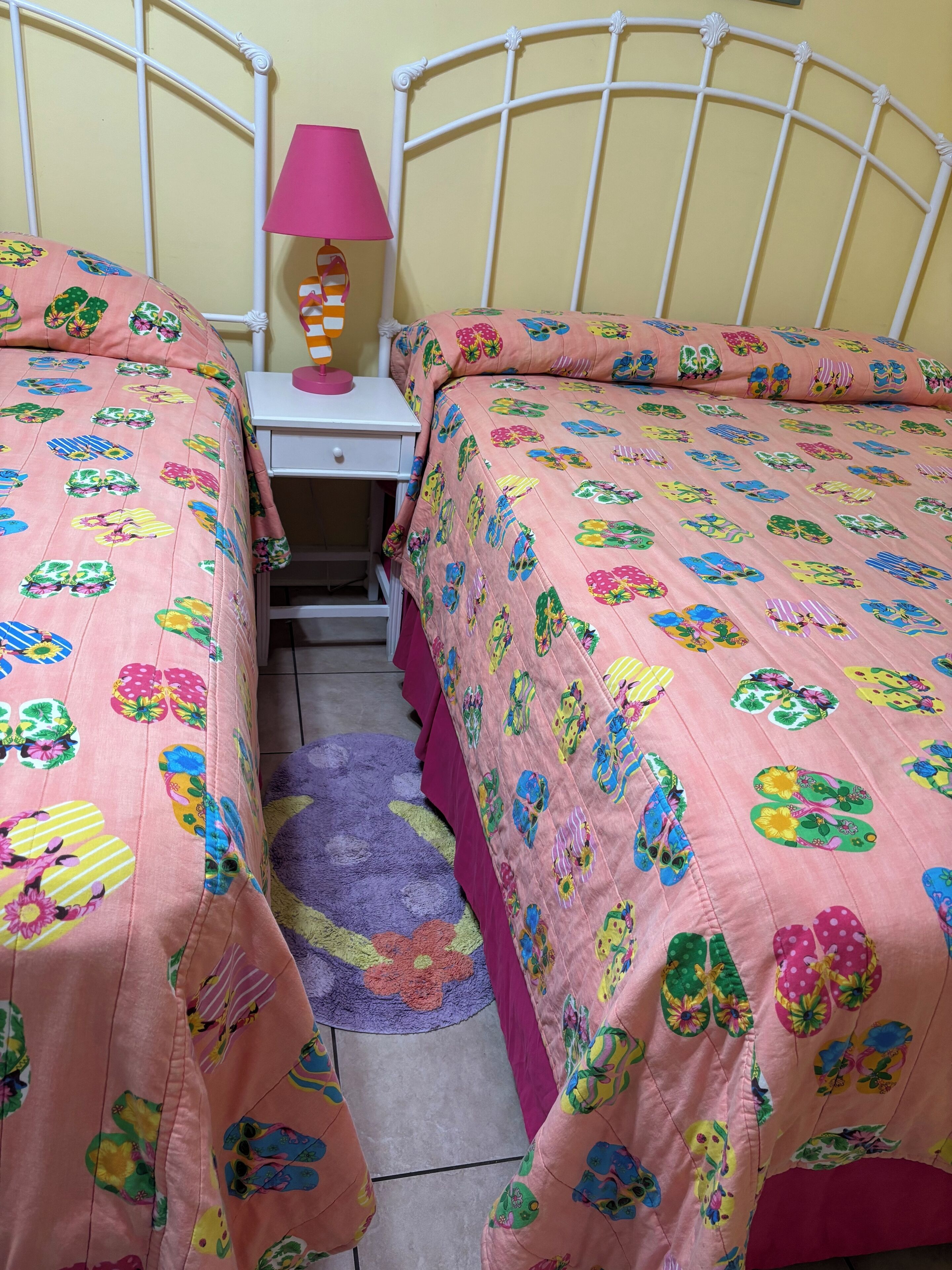 A bedroom with two beds, each with a pink floral bedspread, a white metal headboard, a small bedside table with a lamp, and a purple rug on the floor.