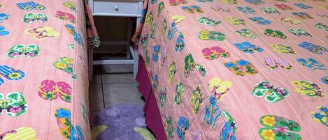 A bedroom with two beds, each with a pink floral bedspread, a white metal headboard, a small bedside table with a lamp, and a purple rug on the floor.