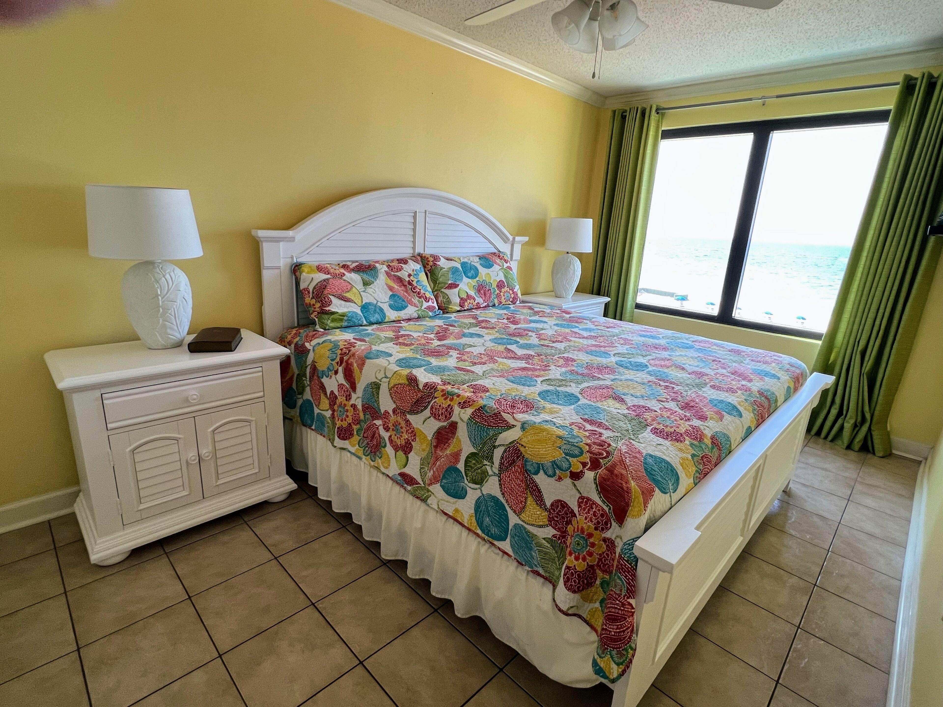 A bedroom with a bed, two nightstands, a ceiling fan, a window with curtains, and a view of the sea.
