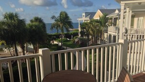 Een balkon met een houten tafel en stoelen, uitkijkend op een verzorgde tuin en het uitzicht op de oceaan.