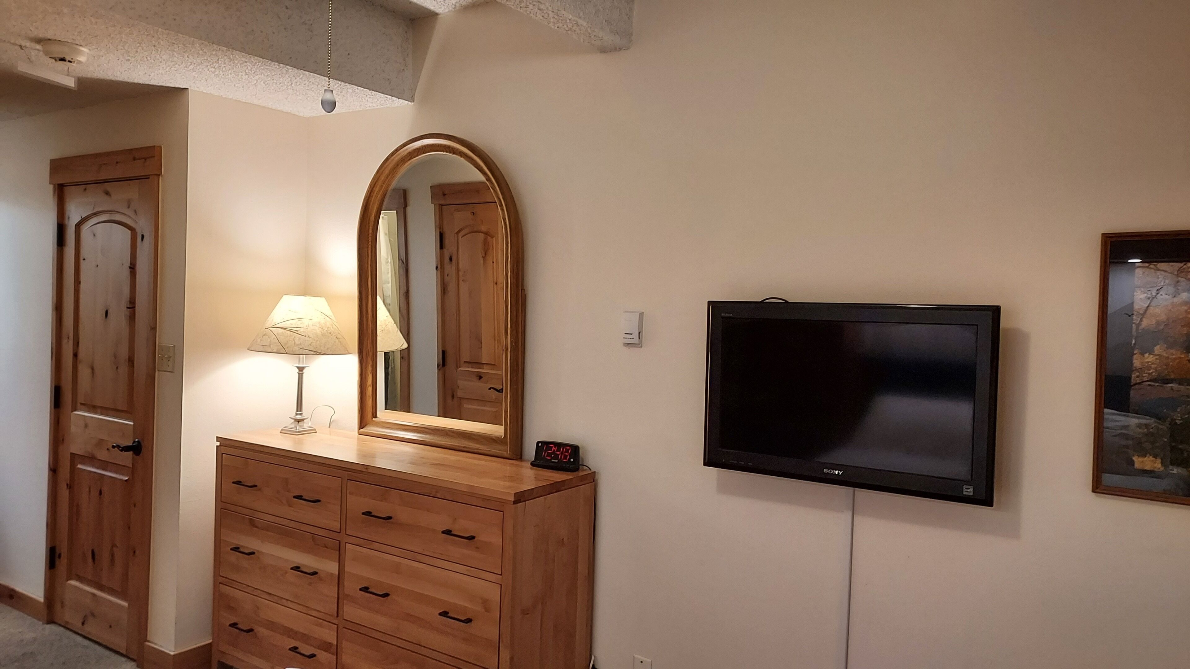 A room with a wooden dresser, a mirror, a lamp, a wall-mounted TV, and a framed picture.