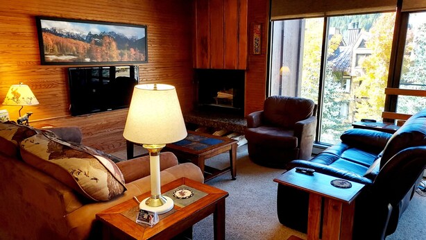 A cozy living room with a fireplace, comfortable seating, a television, and a large window offering a view of trees and buildings. - Ski-In/Ski-out  - 50 ft from SuperBee Lift (Copper Mountain)