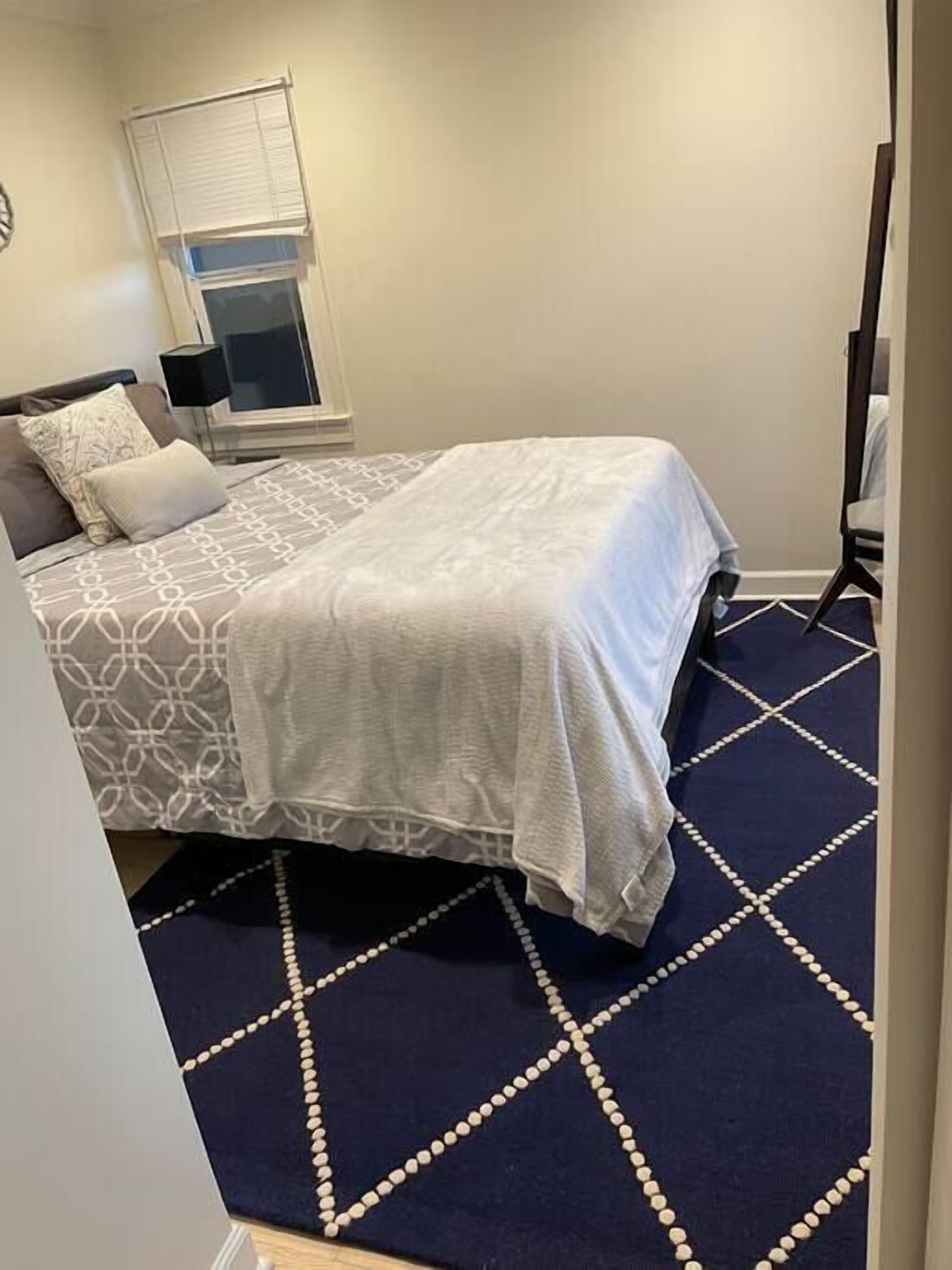 A bedroom with a bed, a window with blinds, a bedside table, and a patterned rug.