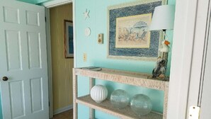 Um quarto com porta branca, prateleira fixada na parede com objetos decorativos, quadro emoldurado e piso ladrilhado.