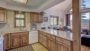 Una cocina con armarios de madera, encimeras de granito y lavavajillas. Hay ventanas que ofrecen luz natural y vistas al exterior.