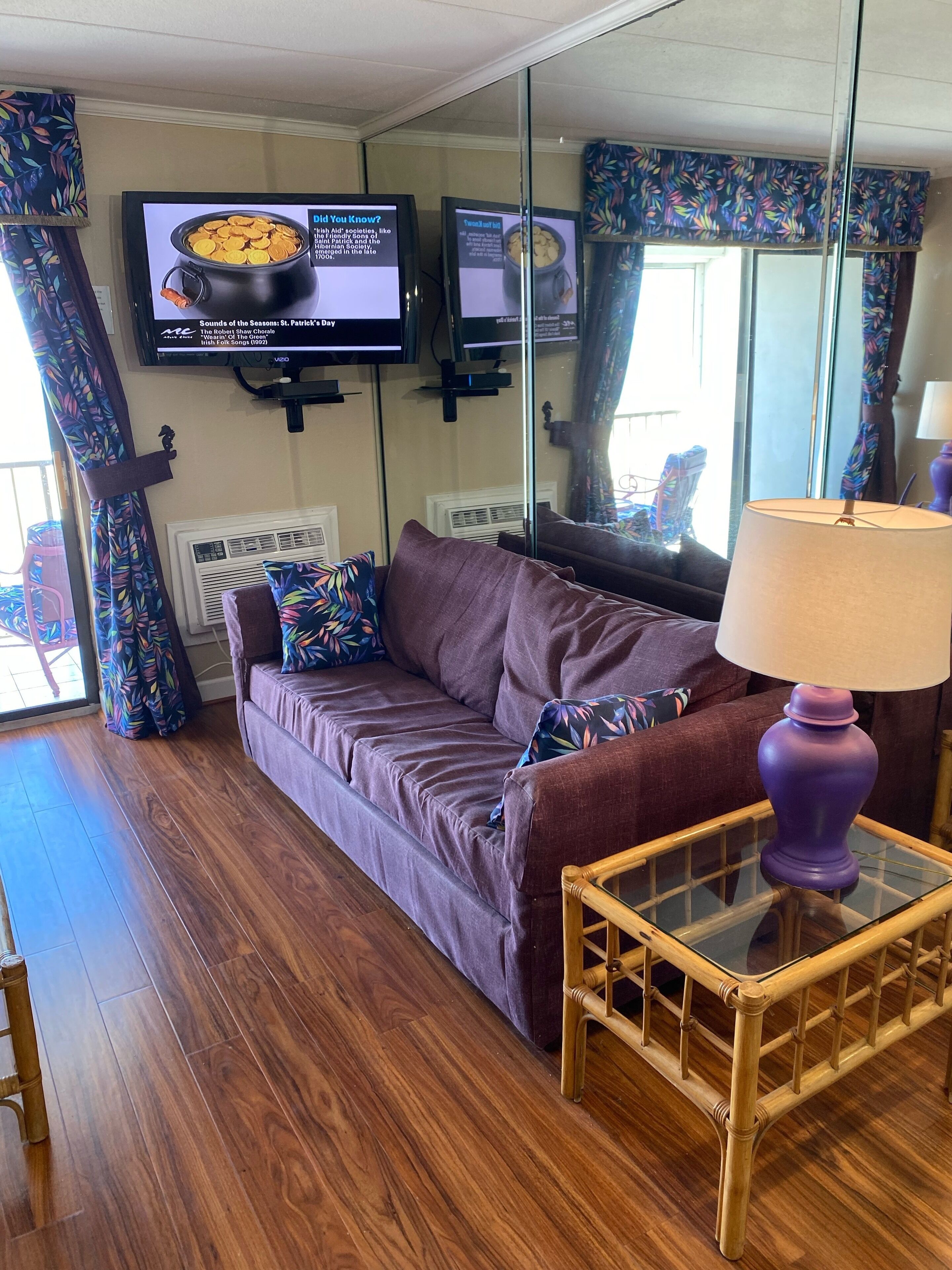 A living room with a sofa, a glass coffee table, a television displaying a program, and a window with curtains.
