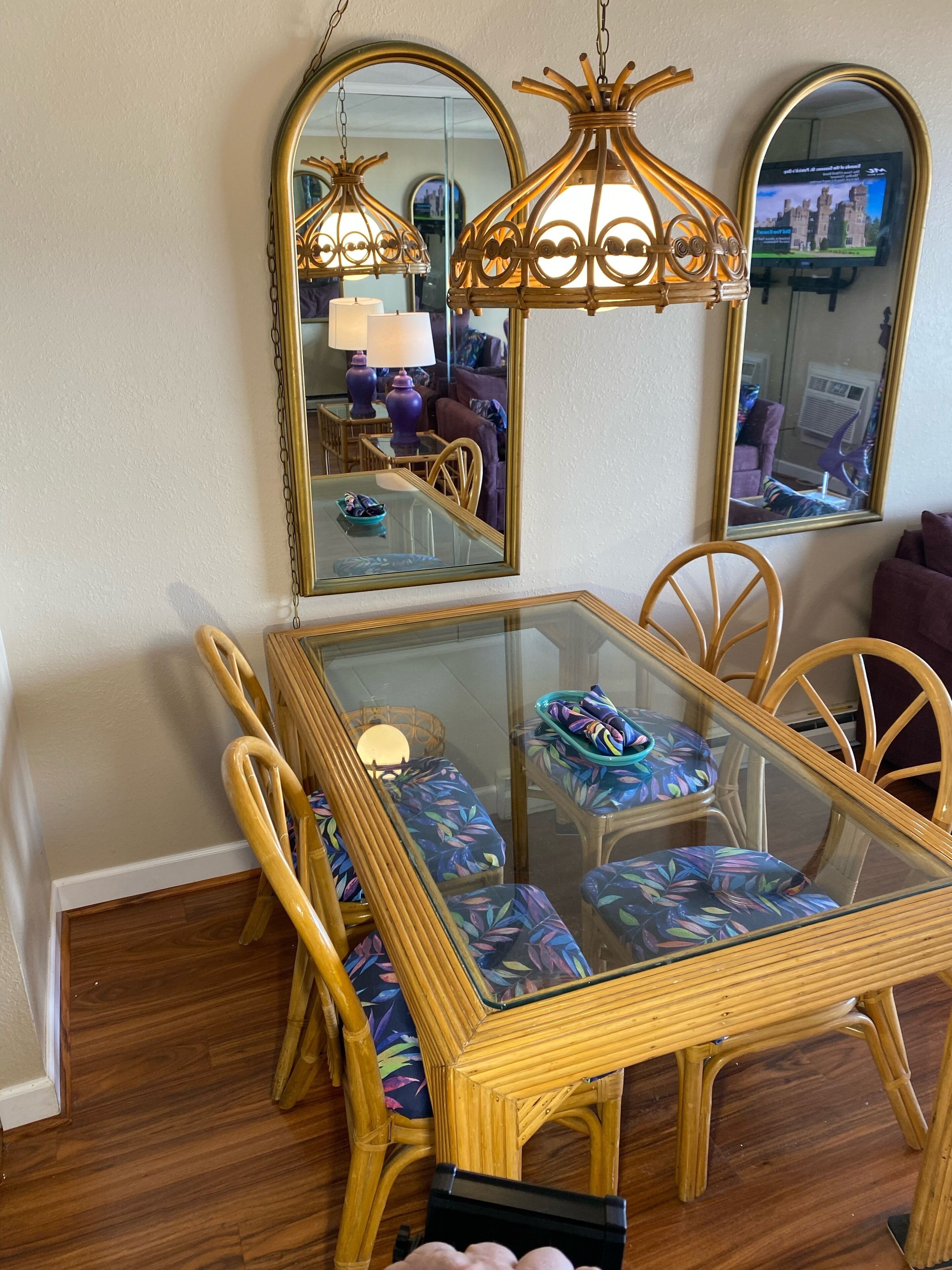 A dining area with a glass-top table, wooden chairs with colorful cushions, and two wall-mounted mirrors.