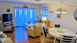 A living room with a TV, sofa, dining table, and a view of the beach through large windows. - AFFORDABLE Escape! OCEAN VIEW, Steps to Beach, Ground Floor 1BR overlooking pool (St. Augustine)