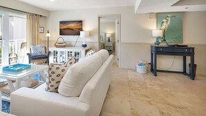 A spacious living room with a white sofa, a coffee table, and a TV mounted on the wall. There is a painting of a bird on the wall, a ceiling fan, and a window with curtains. - Beautiful Ocean Views, Beach Club 337! Steps to beach! (St. Simons Island)