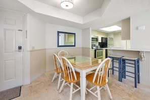 A dining area with a glass table and four chairs, a kitchen in the background, and a white door with a key lock. - Beautiful Ocean Views, Beach Club 337! Steps to beach! (St. Simons Island)
