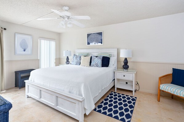 A bedroom with a large bed, a ceiling fan, a bedside table with a lamp, a chair, and a rug. - Beautiful Ocean Views, Beach Club 337! Steps to beach! (St. Simons Island)