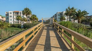 A wooden boardwalk leading to a gated entrance with palm trees and buildings in the background. - Beautiful Ocean Views, Beach Club 337! Steps to beach! (St. Simons Island)