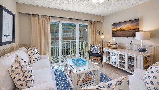 A living room with a sofa, armchair, and a glass-top coffee table. There is a balcony with glass doors and a view of the outside. - Beautiful Ocean Views, Beach Club 337! Steps to beach! (St. Simons Island)