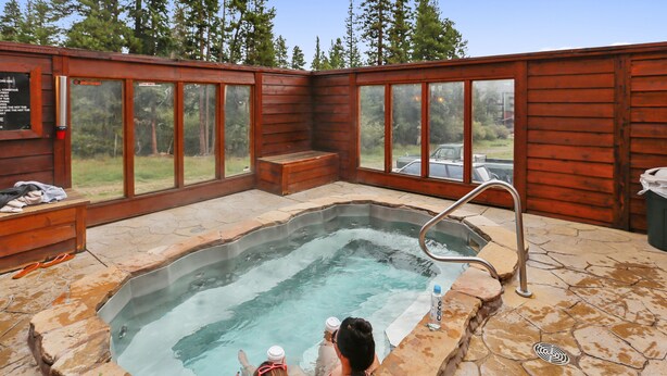 A hot tub with a wooden deck and stone flooring, surrounded by glass windows and a view of trees and a car. - Pine Ridge Ski-In Condo on Four O'Clock Ski Run - 1 block from Downtown (Breckenridge)