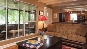 Ein gemütliches Wohnzimmer mit einer Couch, einem Couchtisch mit Büchern und Kerzen, einer roten Lampe und Blick auf einen Essbereich durch große Fenster.
