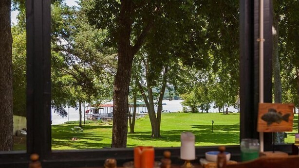 Blick aus einem Restaurant, im Vordergrund ein Tisch und Stühle, im Hintergrund ein Grasland mit Bäumen und einem Gewässer.