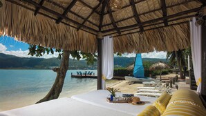A beachside room with a thatched roof, a bed with white linens, a view of the sea, and lounge chairs on the deck. - Beach Front Villa W/Pool/Staff, in Gated Community W/Tennis/Gym (Montego Bay)