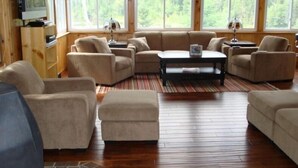 Uma sala de estar espaçosa com sofá de canto, poltronas, mesa de centro e televisão. Janelas amplas oferecem vista para o exterior.