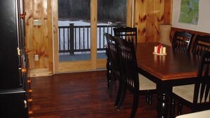 Interior de madeira com mesa e cadeiras de jantar, uma placa com a mensagem 'Welcome to the Woods' e vista para uma paisagem nevada através de uma porta de vidro.