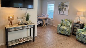 A living room with a TV, a console table with decorative items, two armchairs, a dining area with a table and chairs, and a window with blinds. - The Pool House located in Coveted Old Town with Pool (Port Aransas)