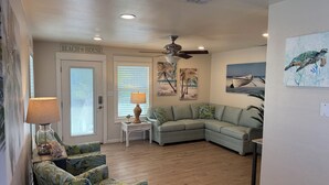 A living room with a sectional sofa, a small side table with a lamp, a ceiling fan, and a door labeled "BEACH HOUSE". - The Pool House located in Coveted Old Town with Pool (Port Aransas)