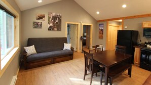 A cozy living room with a dark grey sofa, wooden dining table, and chairs. There is a kitchen area with a black refrigerator and wooden cabinets.