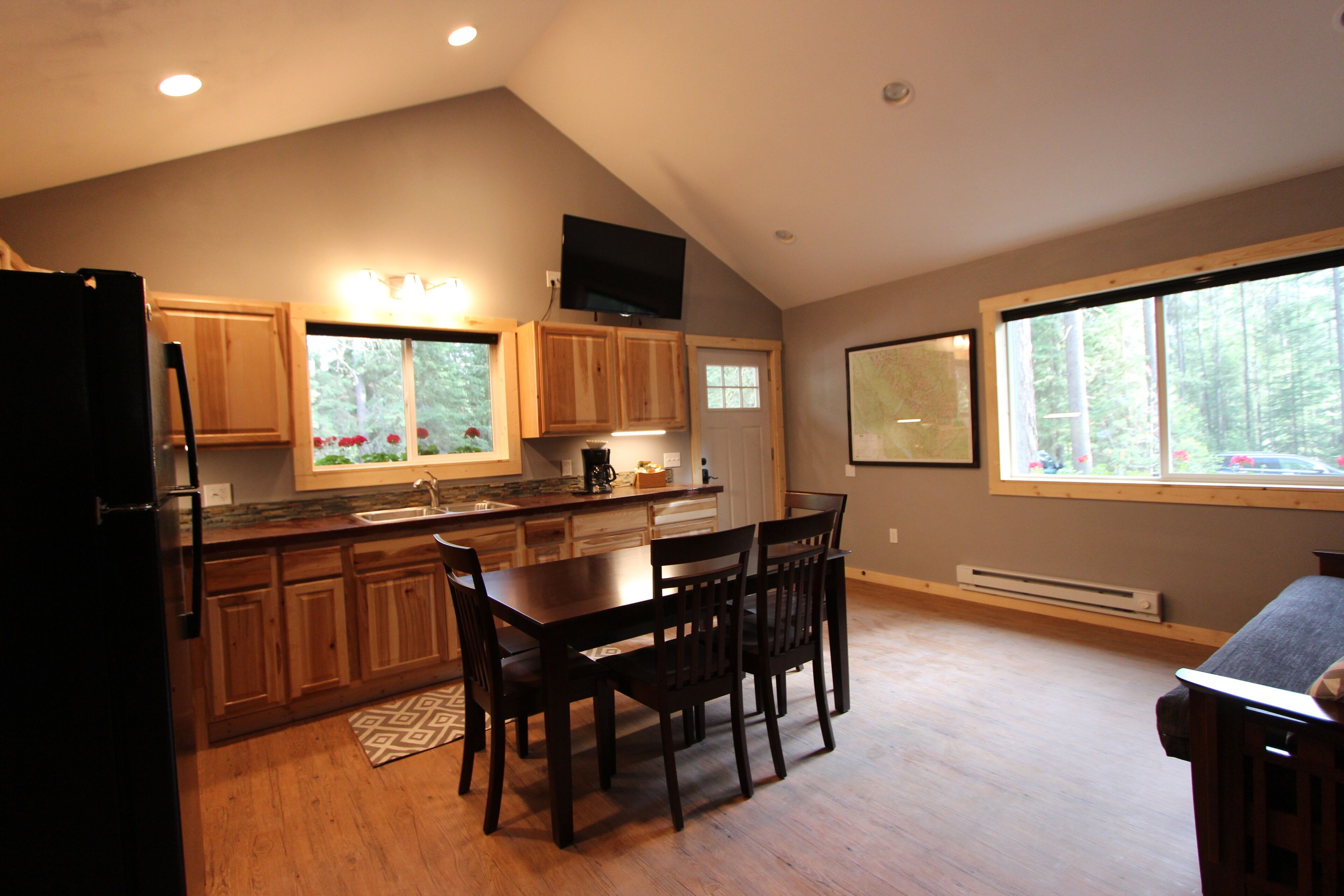 A kitchen with wooden cabinets, a dining table with chairs, a refrigerator, and a window with a view of trees.