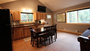 A kitchen with wooden cabinets, a dining table with chairs, a refrigerator, and a window with a view of trees.