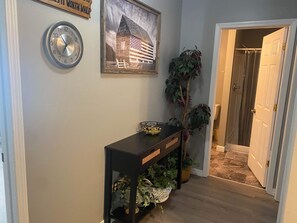 Un couloir avec une console en bois, une horloge, un tableau encadré représentant une grange et une plante en pot.