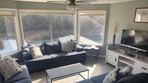 A living room with a grey sofa, a coffee table, and a TV on a stand. There are large windows with a view of trees and a building outside.